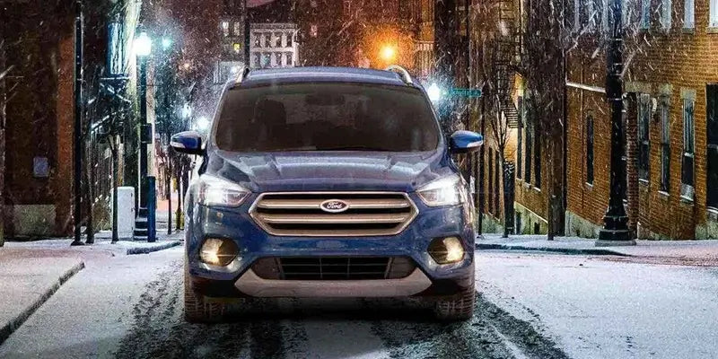 Blue Ford Escape driving through a snowy street at night, showcasing its front view and headlights in a winter setting.