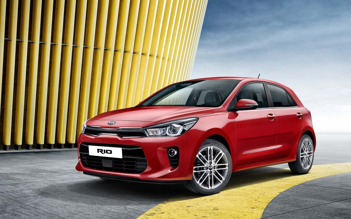 Red Kia Rio parked near modern architecture, showcasing its sleek design and sporty features.